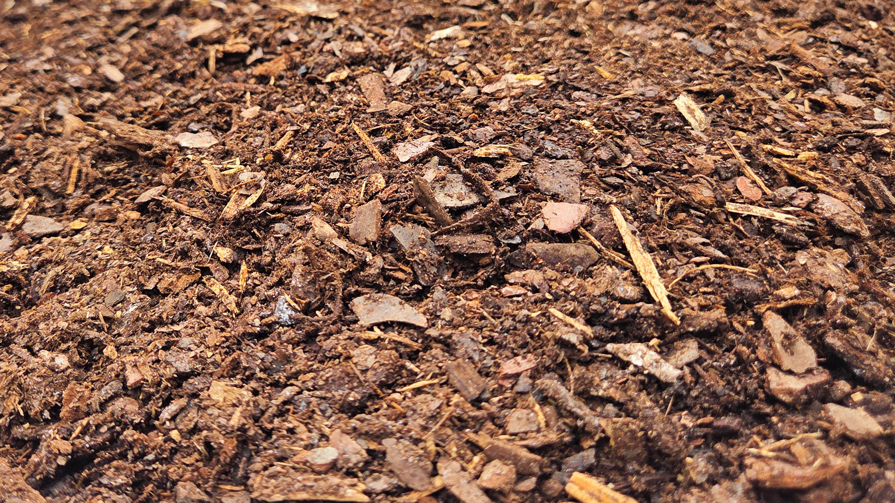 A close-up view of Pine Fines Soil Conditioner with visible wood chips, soil, and organic garden materials behind.